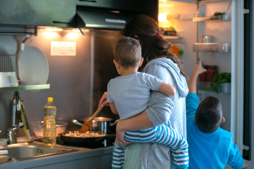 Mother carrying toddler while frying mushrooms for lunch, her older son pointing at stuff in the fridge
